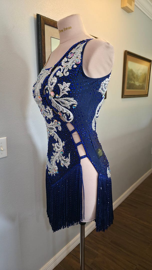 Side view of the Aurora Royale Latin dress in sapphire blue featuring crystal appliqué, pearl embellishments, lattice cutouts, and fringe detailing.