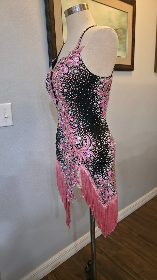 Side front view of the Blush Noir ballroom dress with intricate pink embellishments, crystal accents, and flowing fringe.