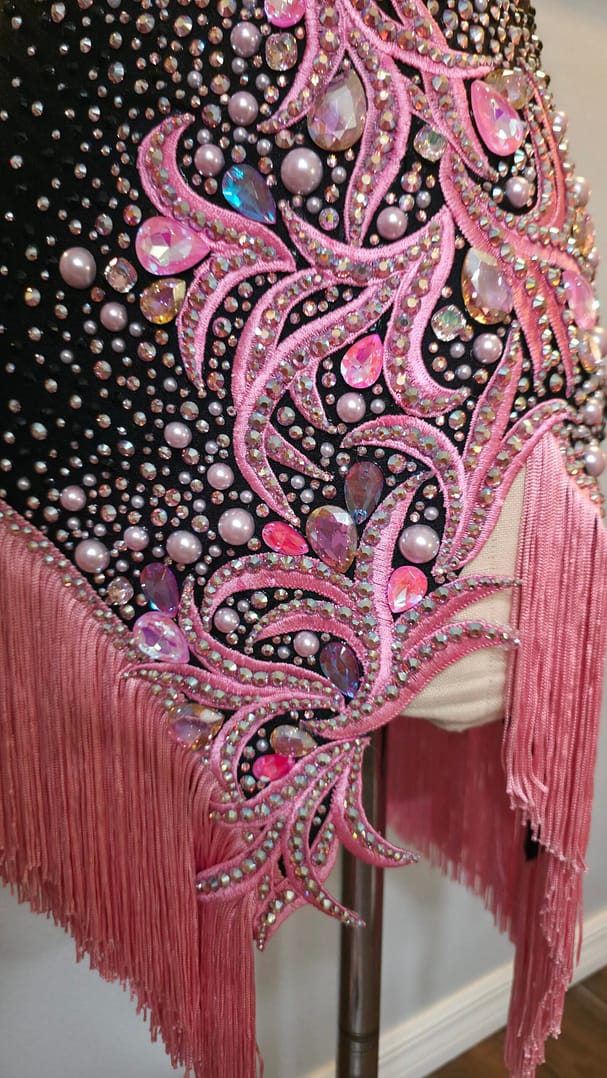 Close-up detail of the Blush Noir Latin dance dress featuring pink crystal appliqué, pearl embellishments, and hand-cut fringe.