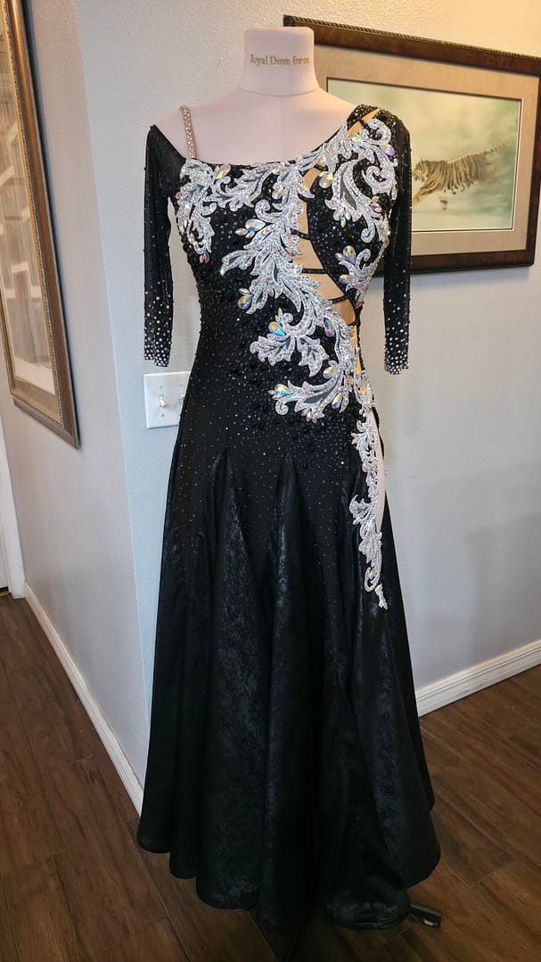 Angled front view of the Crystal Nocturne Signature Series ballroom gown highlighting the flowing black skirt, asymmetrical design, crystal shoulder strap, illusion sleeve, and ornate silver embellishments.