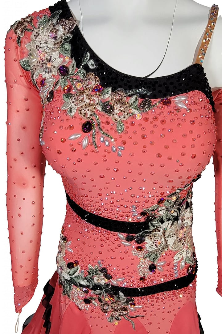 Bruna ballroom dress - detail view — close-up of coral-pink bodice with dense crystals, black sequin neckline, multicolored floral appliqués, and intricate beadwork on sheer mesh sleeve.