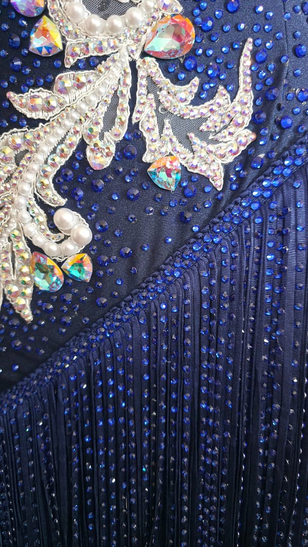 Detailed close-up of sapphire rhinestones and crystal fringe embellishments on the Aurora Royale Latin competition dress.