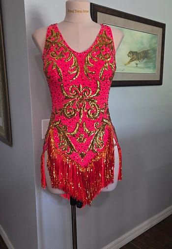 Front view of the Gilded Heat – Performance Series Latin competition dress featuring a neon pink base, ornate gold sequin appliqué, sparkling rhinestones, and asymmetrical fringe.