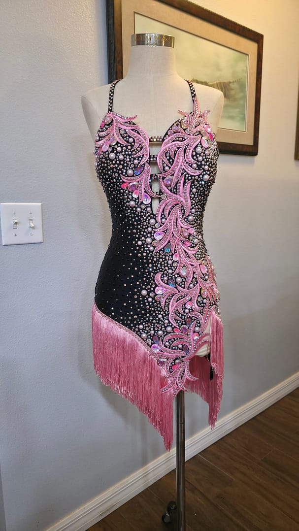 Blush Noir luxury Latin dance dress featuring pink crystal appliqué, pearl embellishments, black base fabric, and flowing fringe accents.