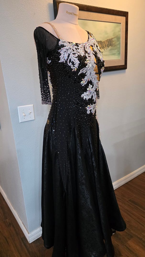 Three-quarter view of the Crystal Nocturne Signature Series black ballroom competition gown featuring a flowing full skirt, illusion sleeve, crystal shoulder strap, and intricate silver appliqué.